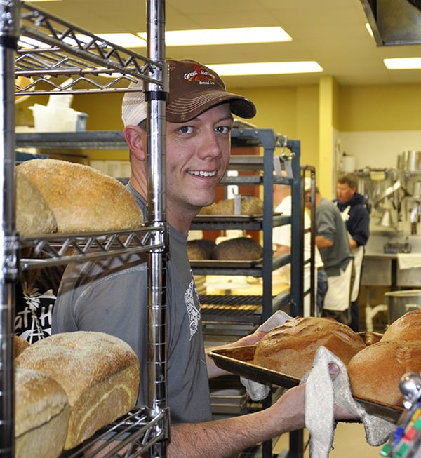 Baker Photo. He is pulling a tray of hot whole wheat bread out of the oven and smiling.