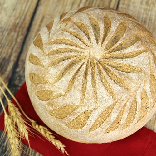 Photo of a round loaf of whole wheat bread on a wood table with a wheat stalk and a red napkin