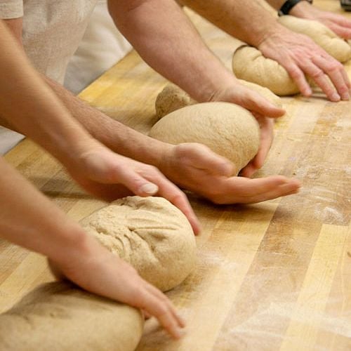 This photo shows hands on a kneading table with loaves of whole wheat dough