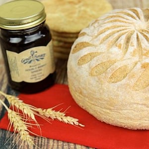 A photo of a honey whole wheat round unsliced loaf, a jar of jam, a stack of cookies, on a red napkin with a wheat stalk